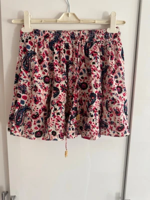 Blommig kortkjol med snörning💕 - Söt kortkjol med blommigt mönster i rött, blått och vitt. Kjolen har resår i midjan och snörning med små pärlor. 