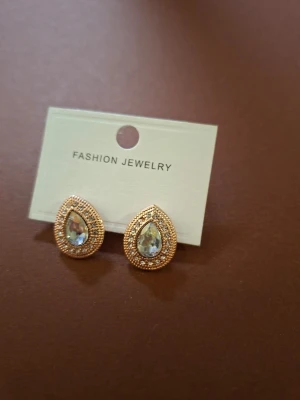 Guldtonade droppformade örhängen - Snygga droppformade örhängen i guldton med glittrande stenar och en större klar sten i mitten. Örhängena har en elegant design med små detaljer runt kanterna som ger extra lyster. Perfekta för att lyfta din stil.