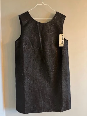 Svart kort klänning med rosett Kappahl - Säljer en svart kort klänning från Kappahl i glansigt, strukturerat material. Klänningen är ärmlös och har en stor dekorativ rosett i ryggen samt djup v-ringning bak. Perfekt för dig som vill sticka ut med en elegant look.