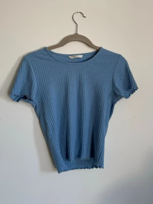 Blå ribbad topp från Only - Säljer en ljusblå ribbad topp från Only med korta ärmar och rund halsringning. Toppen har vågiga kanter vid ärmslut och nederkant för en extra detalj. Perfekt till jeans eller kjol för en enkel och snygg look.