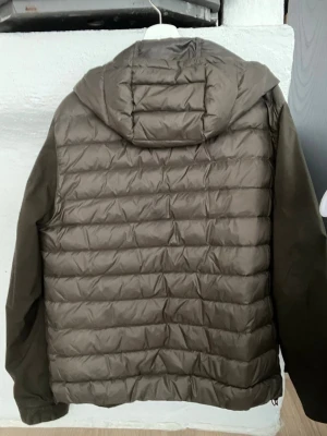 Olivgrön Massimo dutti dunjacka  - Snygg olivgrön dunjacka med quiltad baksida och slät framsida. Jackan har en fast huva, två dragkedjeförsedda fickor framtill och en diskret röd insida vid kragen. Perfekt för kalla vinterdagar och ger en stilren look.