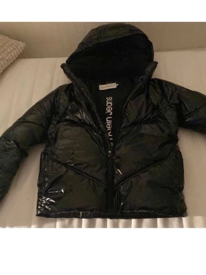 Svart pufferjacka från Calvin Klein - Snygg svart pufferjacka från Calvin Klein Jeans med glansig finish och huva. Jackan har dragkedja framtill och diskreta fickor på sidorna. Perfekt för kalla vinterdagar och ger en cool streetwear-look.