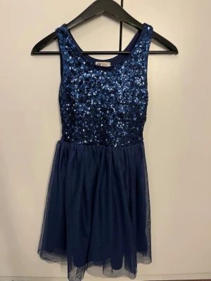 Marinblå paljettklänning H&M - Snygg marinblå kortklänning från H&M med glittrig paljettöverdel och rund halsringning för barn. Kjolen är i tunt, lätt tyg med ett skirt lager ovanpå. 