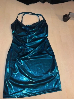 Blå metallic klänning med smala band - Säljer en glansig blå metallic klänning med smala axelband och figurnära passform. Klänningen har en kort längd och är tillverkad i ett syntetmaterial som ger en cool, futuristisk look. Perfekt för dig som vill sticka ut!