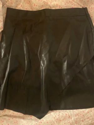 Svart skinnkjol - Snygg svart kortkjol i skinnimitation med omlottdesign och dragkedja i sidan. Kjolen har en stilren look och passar perfekt till en trendig outfit. Materialet ger en cool vibe och den svarta färgen gör den lätt att matcha.