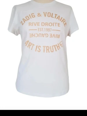 Vit Zadig & Voltaire t-shirt - Vit t-shirt från Zadig & Voltaire med rund hals och korta ärmar. Framsidan har ett tryck i guld med texten 'Zadig & Voltaire', 'Rive Droite', 'Art is Truth' och 'Est. 1997'. Tillverkad i mjuk bomull för en bekväm känsla. 