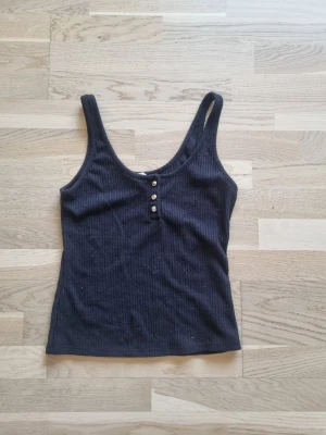 Svart ribbad linnetopp H&M - Snyggt svart ribbat linne från H&M med tre små guldknappar framtill. Toppen har breda axelband och en rundad urringning. Väldigt skönt material. 