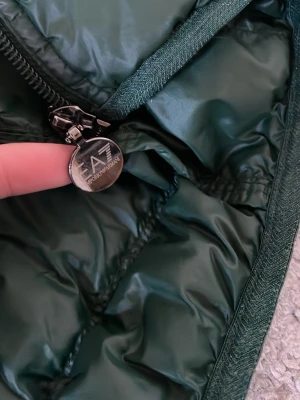 Mörkgrön pufferjacka från Armani - Snygg mörkgrön pufferjacka från Emporio Armani med glansigt yttertyg och quiltad design. Jackan har dragkedja med EA7-logga och praktiska sidofickor. Perfekt för kalla dagar när du vill hålla stilen.