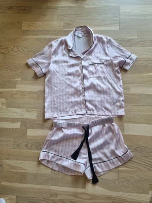 Rosa randig pyjamas från Gina Tricot - Söt pyjamas från Gina Tricot i glansigt, ljusrosa tyg med vita ränder. Setet består av en kortärmad skjorta med knappstängning och bröstficka samt matchande shorts med svart dragsko och svarta kantdetaljer. Perfekt för mysiga kvällar hemma.