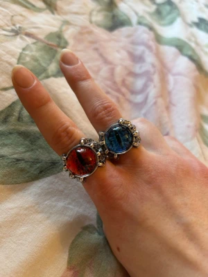 Två chunky ringar med färgade stenar - Säljer två chunky ringar med stora, runda stenar i rött och blått. Ringarna har en silverfärgad metallbas med detaljer runt stenen som ger en cool och unik look. Perfekta för dig som vill sticka ut och addera färg till din stil. Ringarna är justerbara i storlek. 