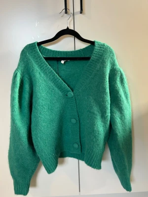 Grön fluffig kofta med v-ringning - Mysig grön kofta med v-ringning och stora knappar framtill. Koftan har en fluffig, stickad känsla och långa ärmar med lätt puff. Ribbstickade muddar vid ärmslut och nederkant. Perfekt för lager på lager och en chill look.