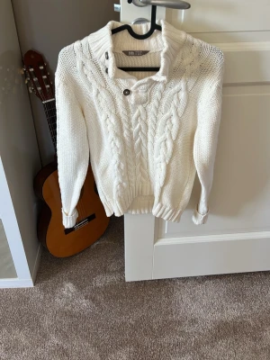Vit kabelstickad tröja Kids by Lindex - Säljer en vit kabelstickad tröja från Kids by Lindex med hög krage och två dekorativa knappar vid halsen. Tröjan har långa ärmar, ribbade muddar och ett klassiskt flätmönster framtill. Perfekt för kyliga dagar och riktigt mysig.