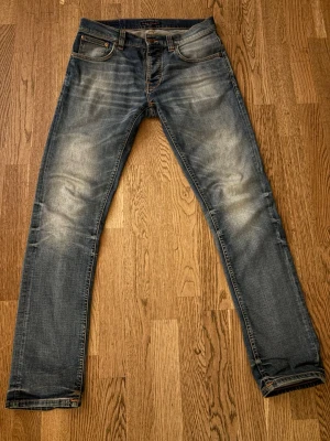 Nudie jeans Grim Tim  - Säljer ett par blå slim fit jeans från Nudie Jeans med klassiska bakfickor och snygga slitningar på knän och lår. Jeansen har en smal passform och är tillverkade i mjuk denim med orange sömmar. Perfekta för en avslappnad och trendig look.