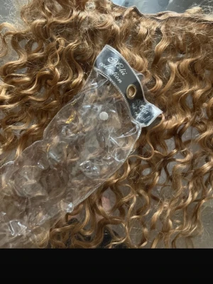 Blonda lockiga löshår från Chivi - Säljer ett paket blonda äkta löshår från Chivi collection. Håret är långt, har tighta lockar och en naturlig glans. Perfekt för dig som vill ha extra volym och längd med en cool, lockig look. Fästet är insytt och passar till clips eller insytt hår.