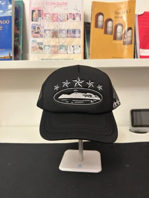 Svart truckerkeps med broderade stjärnor - Snygg svart truckerkeps med mesh på sidorna och bak. Framtill finns ett broderat motiv av ett skepp och fem stjärnor i vitt. På sidan är fler stjärnor broderade. Justerbar passform med klassisk böjd skärm. Perfekt för en streetwear-look.