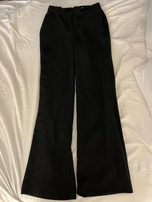 Svarta vida kostymbyxor - Säljer ett par svarta kostymbyxor med vida ben och hög midja. Byxorna har resår baktill i midjan, bälteshällor och två fuskfickor bak. Klassisk och stilren look som passar till många olika outfits.