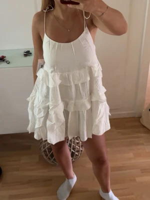 Vit volangklänning med tunna band - Supersöt vit kortklänning med tunna axelband och flera lager volanger. Klänningen har öppen rygg och en luftig passform som känns perfekt för varma dagar. Materialet är lätt och känns som bomull. Perfekt för sommarens alla tillfällen!