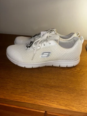 Helt nya vita mesh sneakers från Skechers - Helt nya snygga vita sneakers från Skechers med mesh-överdel och klassisk logga på sidan. Skorna har vita snören och en lätt, flexibel sula i vitt och grått. Perfekta för dig som gillar sportig och clean stil. Original pris 1300kr