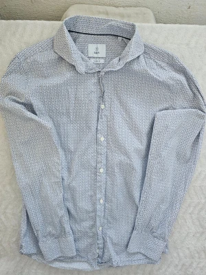 Vit och blå mönstrad skjorta från Bläck - Snygg vit skjorta från Bläck med småblått mönster över hela tyget. Skjortan har klassisk krage, knappar framtill och långa ärmar. Perfekt för dig som gillar stilrena detaljer och en modern passform.