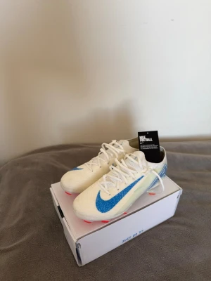 Nike vita fotbollsskor med blå swoosh - Snygga vita fotbollsskor från Nike med en stor blå swoosh på sidan och detaljer i ljusblått. Skorna har snörning och dobbar i neonrött för bra grepp på planen. Tillverkade i syntetmaterial som ger lätt känsla och bra bollkontroll. Perfekta för dig som vill sticka ut på planen. Använda en gång, pris kan diskuteras.