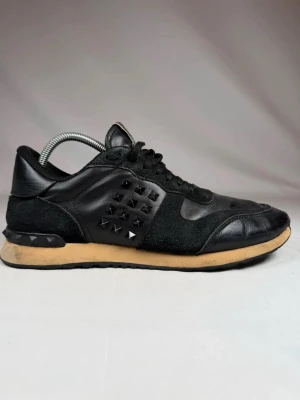 Svarta Valentino Rockrunners  - Snygga svarta sneakers från Valentino med coola pyramidformade nitar på sidorna. Skorna är gjorda i en mix av läder och mocka, har svarta skosnören och en beige sula som ger en schysst kontrast. Perfekta för dig som vill sticka ut med en edgy stil.