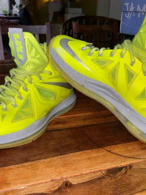 Nike LeBron X gula high sneakers - Nike LeBron X sneakers i neon-gult med silverfärgade swoosh-loggor och detaljer. Skorna har högt skaft, meshpaneler för extra ventilation och en robust sula i grått och genomskinligt gummi. Perfekt för dig som gillar att sticka ut med sportig vibe.