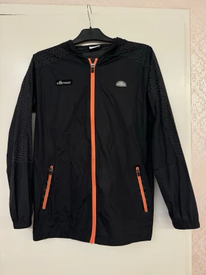 Vindjacka från Ellesse - Snygg vindjacka från Ellesse med orange dragkedjor och detaljer. Jackan har huva, två fickor med dragkedja och diskret mönster på axlar och ärmar. Tillverkad i lätt och vindtätt material, perfekt för blåsiga dagar.