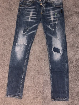 Blå slitna jeans från Dsquared2 - Säljer ett par blå jeans från Dsquared2 med slitningar och hål på båda benen. Jeansen har en tvättad look med ljusa partier och detaljer som ger en riktigt trendig och avslappnad vibe. Klassisk femficksmodell och knappgylf. Perfekta för dig som gillar streetstyle.