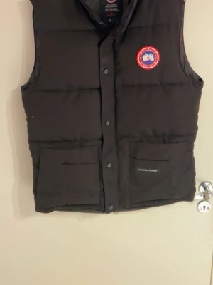 Svart dunväst från Canada Goose - Säljer en svart dunväst från Canada Goose med klassisk logga på bröstet och två stora fickor framtill. Västen har tryckknappar och hög krage, perfekt för kyliga dagar. Tidlös design och riktigt skön passform.