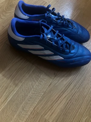 Blå Adidas Copa elite fotbollsskor - Säljer ett par blå Adidas Copa fotbollsskor med klassisk design och vita ränder på sidorna. Skorna har snörning och är tillverkade i syntetmaterial med en låg profil för bra bollkontroll. Perfekta för dig som vill ha stilrena och bekväma skor på planen.