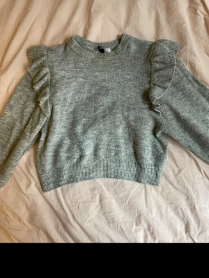 Grå stickad tröja med volanger - Mysig grå stickad tröja med volangdetaljer längs axlarna och långa ärmar. Jag har fått den av en vän men den har tyvärr inte kommit till användning för mig