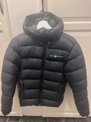 Svart dunjacka från Sail Racing - Snygg svart dunjacka från Sail Racing med huva och logga på bröstet samt en cool detalj på huvan. Jackan har en puffig design med breda kanaler och dragkedja framtill. Perfekt för kalla vinterdagar och riktigt varm.