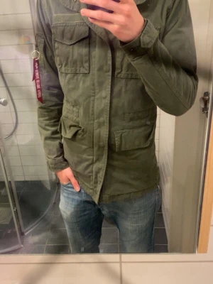 Militär Jacka Alpha Industries - Säljer en olivgrön parkajacka med flera stora fickor framtill och klassisk krage. Jackan har långärmad design och stängs med både knappar och dragkedja. Perfekt för dig som gillar en avslappnad och funktionell stil.