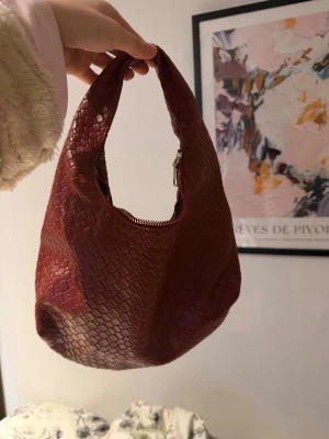 Röd handväska med ormmönster - Cool röd handväska med ormmönstrad yta och rundad form. Väskan har ett kort handtag och en dragkedja upptill. Materialet ser ut som konstläder med reptilstruktur, perfekt för att ge din outfit en edgy touch.