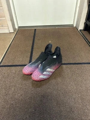 Adidas Predator rosa/svarta fotbollsskor - Säljer ett par Adidas Predator fotbollsskor utan snörning i en snygg färgkombination av svart och rosa med vita detaljer. Skorna har en strumpliknande passform och är tillverkade i syntetmaterial för bra bollkontroll och komfort på planen.
