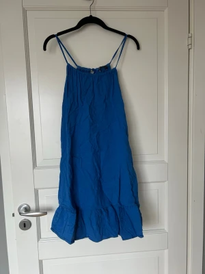 Blå ärmlös sommarklänning - Säljer en blå, ärmlös klänning med smala axelband och volang nedtill. Klänningen har en lös passform och är tillverkad i ett lätt tyg, perfekt för varma dagar. Enkel design med liten knytning framtill vid halsen.