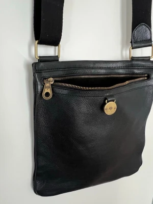 Svart axelväska i skinn från Mulberry - Snygg svart axelväska i äkta skinn från Mulberry med justerbar axelrem i tyg. Väskan har en framficka med dragkedja och guldfärgade metalldetaljer, inklusive Mulberrys klassiska logga på knappen. Perfekt för dig som gillar stilrena accessoarer.