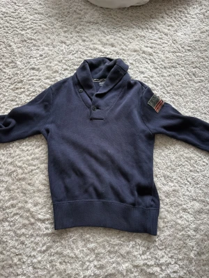  stickad tröja från Ralph lauren  - Mörkblå stickad tröja från Polo Jeans Company by Ralph Lauren med sjalkrage och två knappar vid halsen. Tröjan har ribbade muddar och en amerikansk flagga som patch på ena ärmen. Perfekt för dig som gillar klassisk och avslappnad stil. Skriv vid funderingar eller frågor✌🏼
