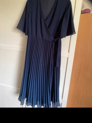 Marinblå omlottklänning med plisserad kjol - Snygg marinblå omlottklänning med v-ringning och knyt i midjan. Kjolen är plisserad och klänningen har korta, vida ärmar. Perfekt för dig som gillar en elegant och stilren look.