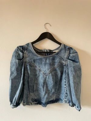 Blå jeansblus med puffärm  - Trendig blå jeansblus från Lindex  med puffiga långärmar och rund halsringning. Blusen har en markerad söm framtill och dragkedja i ryggen för en cool look. Tillverkad i denim med en croppad passform som ger en modern vibe.