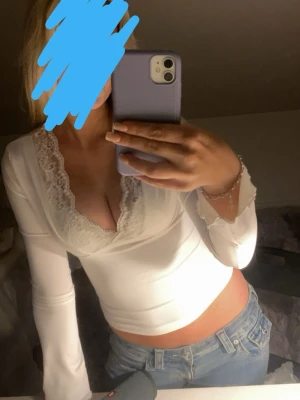 Vit långärmad topp med spets - Säljer en vit långärmad topp med djup v-ringning och fina spetsdetaljer runt halsen. Toppen är figurnära och har en croppad passform som passar perfekt till jeans eller kjol. Superfin och enkel att styla för en trendig look.