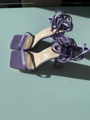 Lila pumps med snörning och fyrkantig tå - Säljer ett par trendiga lila pumps med fyrkantig tå och höga blockklackar. Skorna har tunna remmar över tårna och långa snören som lindas runt ankeln. Tillverkade i ett slätt material som ger en stilren look. Perfekta för dig som vill sticka ut!