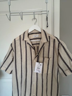 Beige randig skjorta Pull&Bear - Säljer en beige skjorta från Pull&Bear med svarta vertikala ränder. Skjortan har korta ärmar, klassisk krage, bröstficka och knäppning framtill. Tillverkad i ett luftigt material som passar perfekt till varmare dagar.