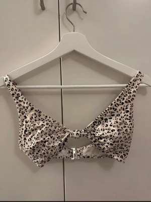 Leopardmönstrad bikiniöverdel HM - Snygg bikiniöverdel i leopardmönster med vit bas och svarta detaljer. Modellen har en dekorativ knut framtill och metallspänne i ryggen. Justerbara axelband för bästa passform. Perfekt för strandhäng eller poolparty.
