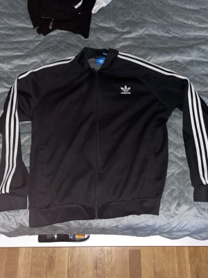 Svart Adidas hoodie med dragkedja - Svart hoodie från Adidas med klassiska vita ränder längs ärmarna och logotyp på bröstet. Tröjan har dragkedja framtill och ribbade muddar. Perfekt för en sportig och avslappnad look.