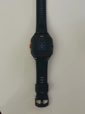 Svart smartklocka med silikonarmband - Snygg svart smartklocka med fyrkantig display och silikonarmband. Klockan har flera knappar på sidorna och färgdetaljer i orange. Perfekt för dig som vill ha koll på notiser, träning och hälsa direkt på handleden. Modern design och bekväm passform.