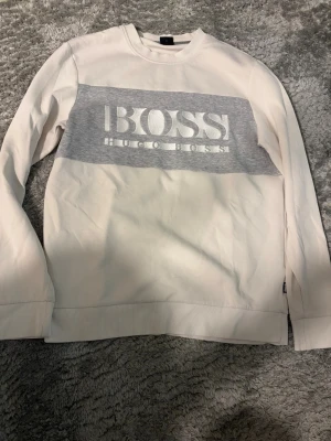 Vit sweatshirt från Hugo Boss - Snygg vit sweatshirt från Hugo Boss med grå panel och stor logga framtill. Tröjan har rund halsringning, långa ärmar och ribbade muddar. Perfekt för en clean och stilren look.