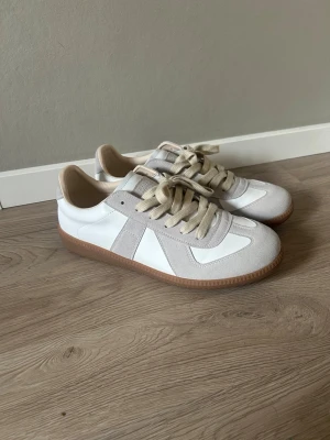 Maison margiela sneakers storlek 43 - Storlek 43 använda bara ett par gånger och jag säljer för att dom passar tyvärr inte mig längre. Snygga sneakers i vitt och beige med ovandel i läder och mocka, samt beige snören och gummisula. Klassisk låg modell som passar till många olika stilar. Perfekta för dig som gillar stilrena och tidlösa skor.