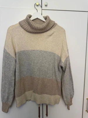 Beige och grå polotröja Solutions! - Mysig stickad polotröja från Solutions! i färgblock med beige, ljusgrå och ljusbrun. Tröjan har bred polokrage, långa ärmar och ribbade muddar. Perfekt för kalla dagar och enkel att matcha med jeans eller kjol.