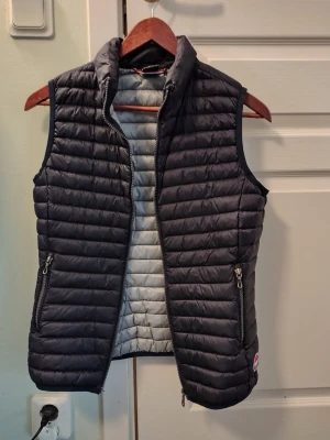 Marinblå dunväst med fickor - Snygg marinblå dunväst med  krage. Västen har två praktiska fickor med dragkedja och en diskret logga nertill på ryggen. Insidan är ljusgrå och materialet är lätt och smidigt, perfekt för lager-på-lager-stil. Passar storlek xxs- xs